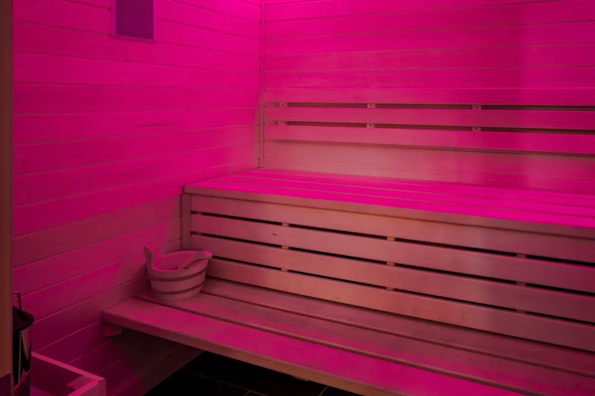 An inviting interior sauna is bathed in calming pink light, featuring wooden benches and a bucket for traditional sauna experiences. The ambiance is both soothing and restorative, suggesting a dedicated space for relaxation and wellness rituals.