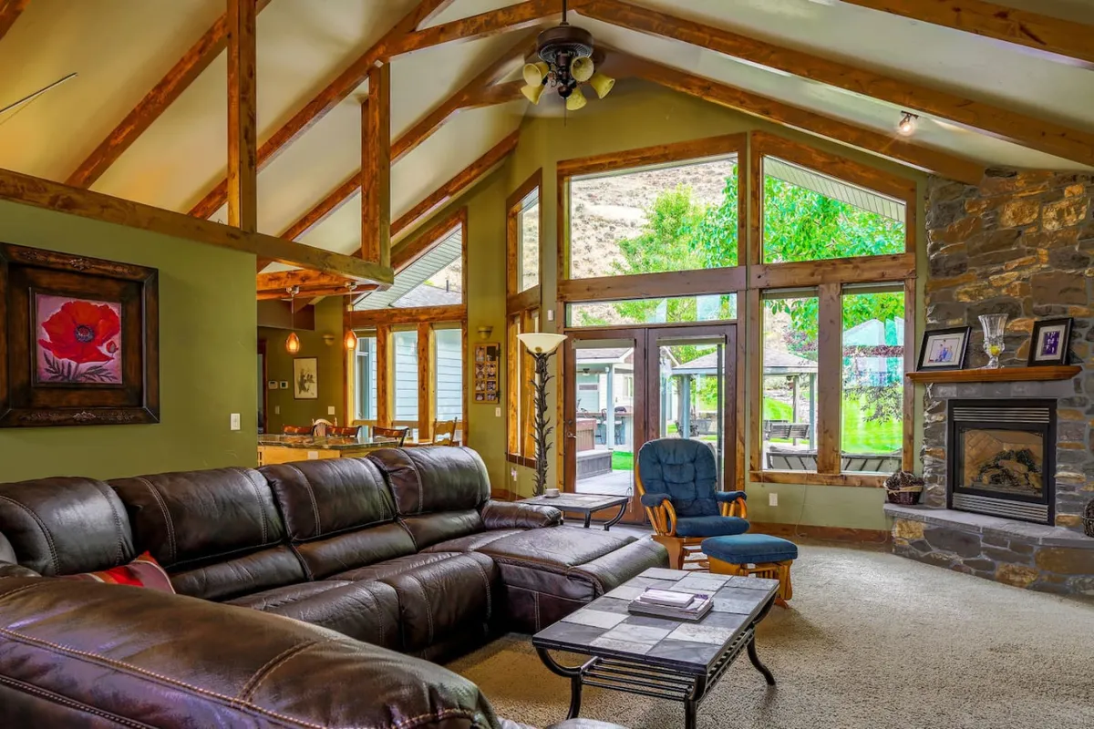 A spacious living room featuring a large leather sectional sofa facing a rustic stone fireplace. Sunlight streams through expansive windows, revealing scenic views. The ceiling is accented with heavy wooden beams, enhancing the room's welcoming and cozy atmosphere.