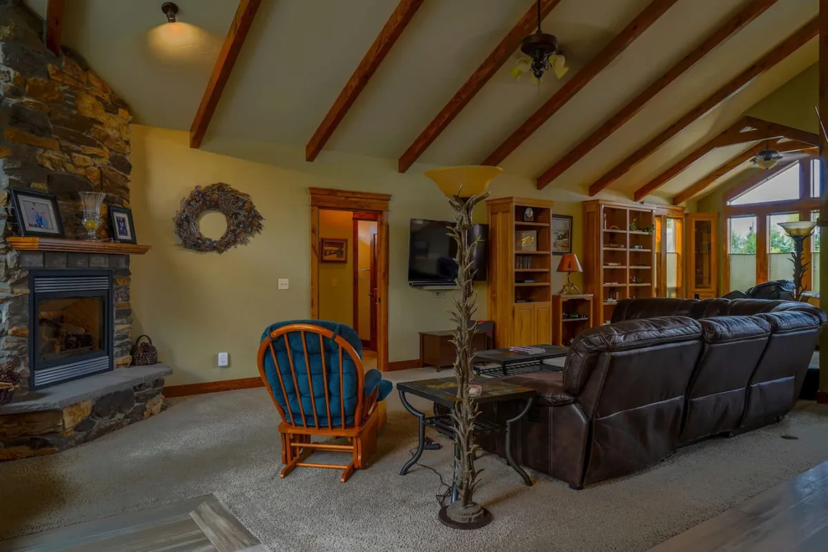 A warm living area anchored by a majestic stone fireplace, comfortable seating, exposed wooden beams, and expansive windows framing scenic outdoor views. The layout is open and perfect for gatherings or peaceful fireside moments.