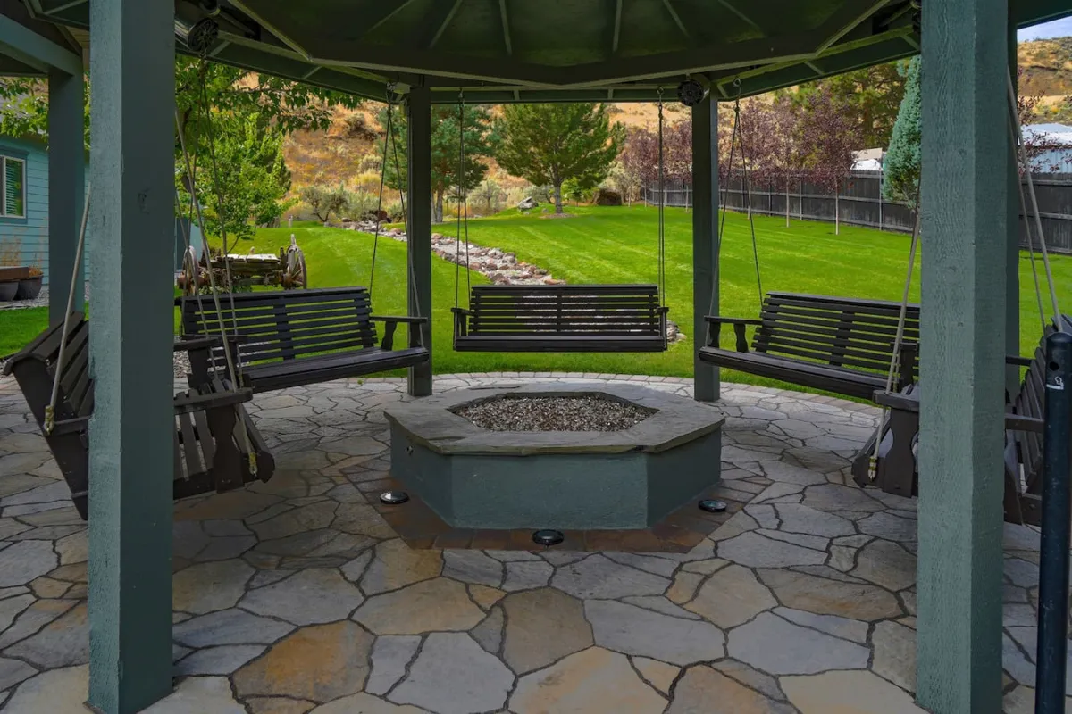 A cozy outdoor living area featuring swing seats circling a fire pit, set on a stone patio surrounded by a lush green lawn. The space is ideal for evening gatherings, roasting s'mores, and sharing stories under the stars.