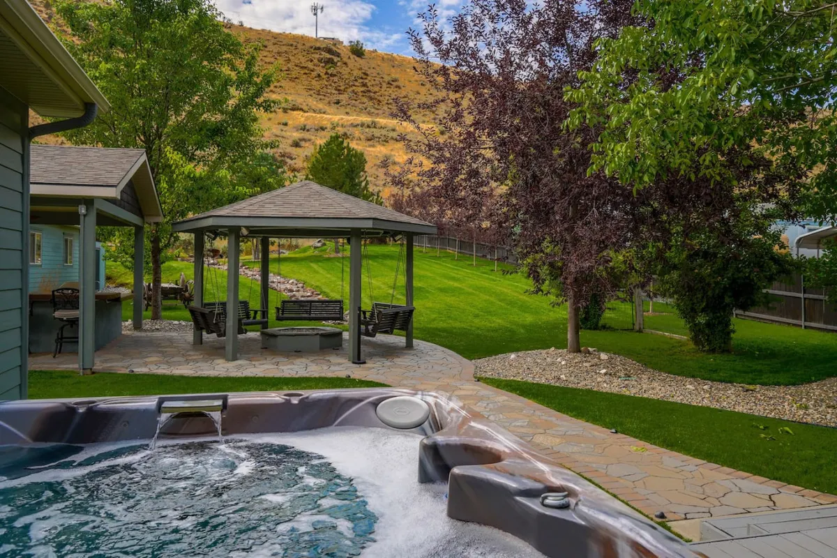 This exterior image showcases a hot tub and gazebo set amidst a lush green lawn and mature trees. The tranquil setting features inviting spaces for both relaxation and social gatherings, with the hot tub perfectly positioned for evening stargazing and the gazebo ideal for sunset views or morning yoga.