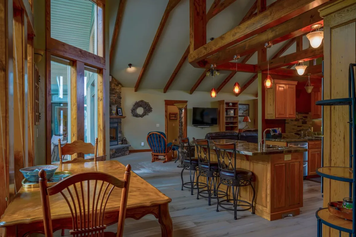 An open-plan dining and living area showcasing a substantial wooden dining table surrounded by chairs and bar stools. The adjoining living space offers comfortable seating, kitchen cabinetry, and is flooded with natural light from large windows.